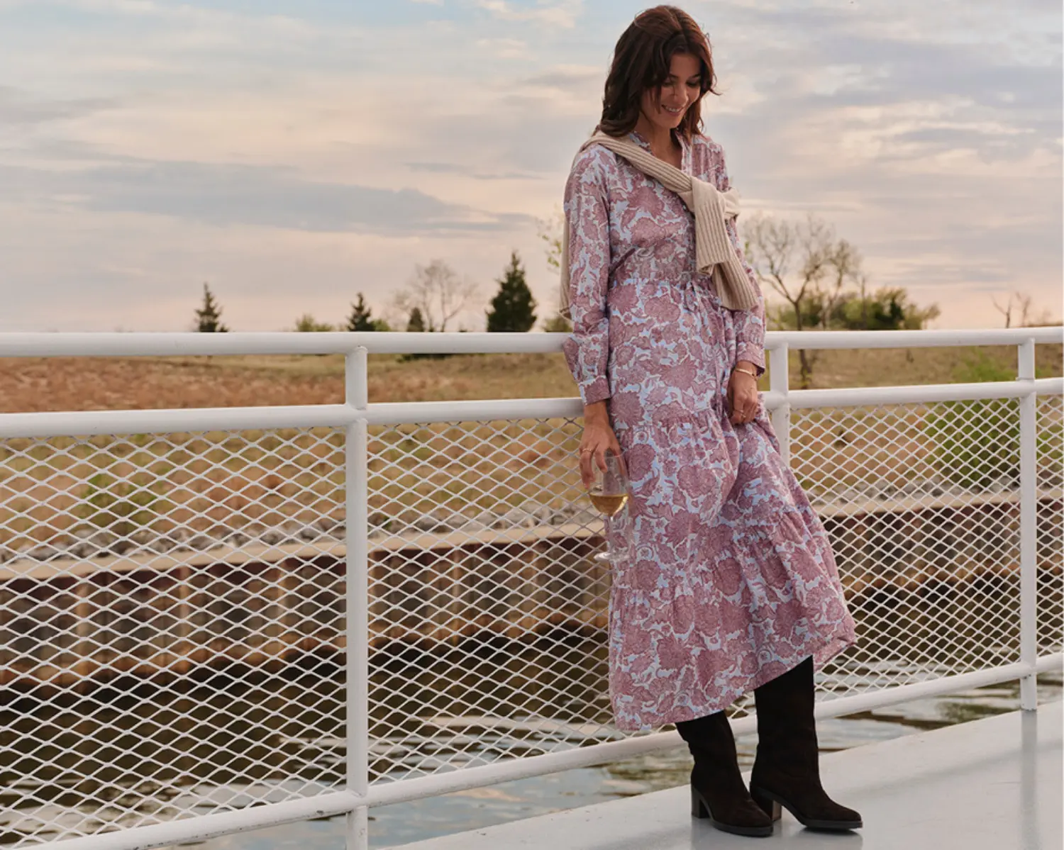 Woman on a boat wearing a full length floral dress with a creme sweater and brown boots