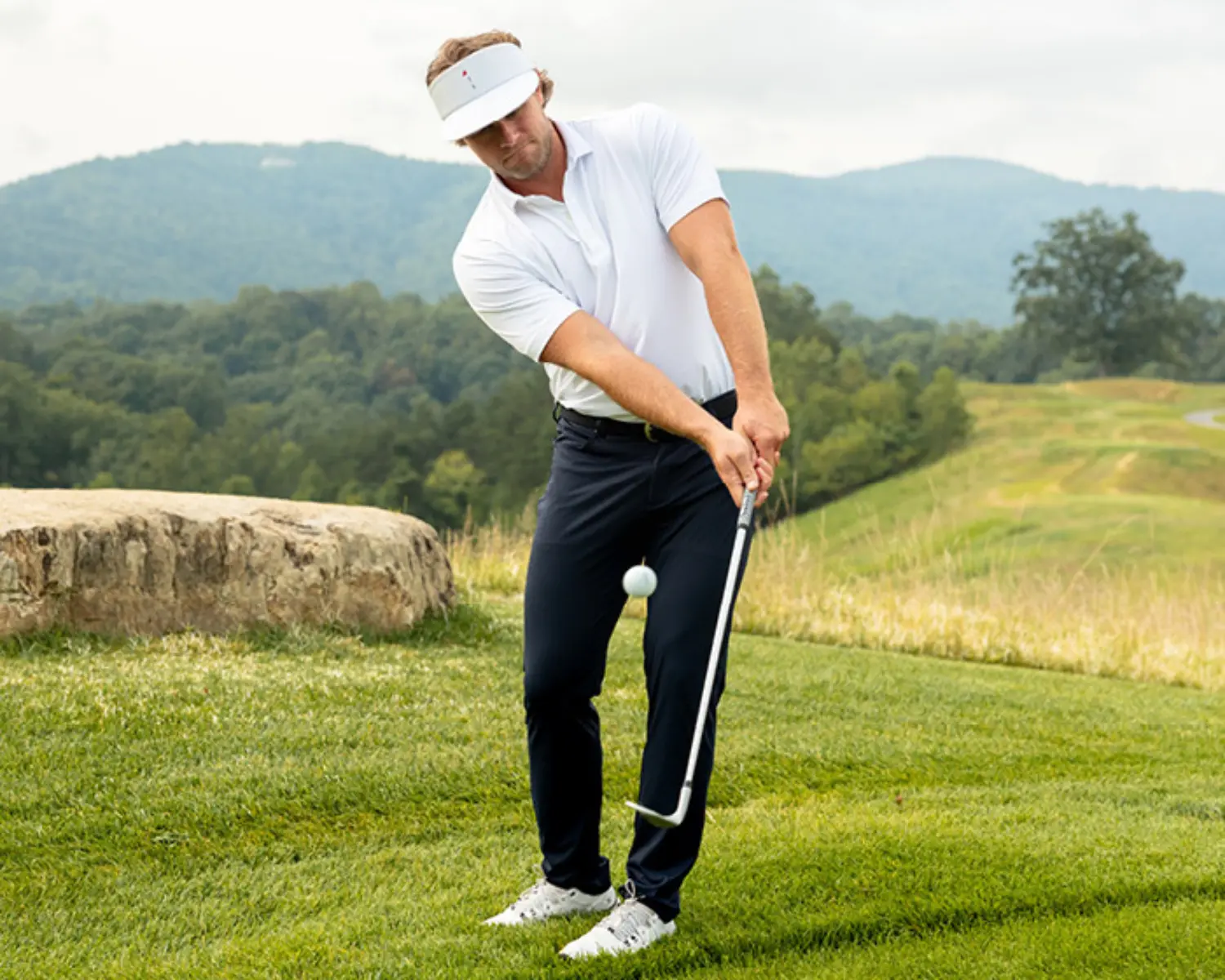 Man dressed in a performance polo, golf visor, and golf pants playing golf in the mountains