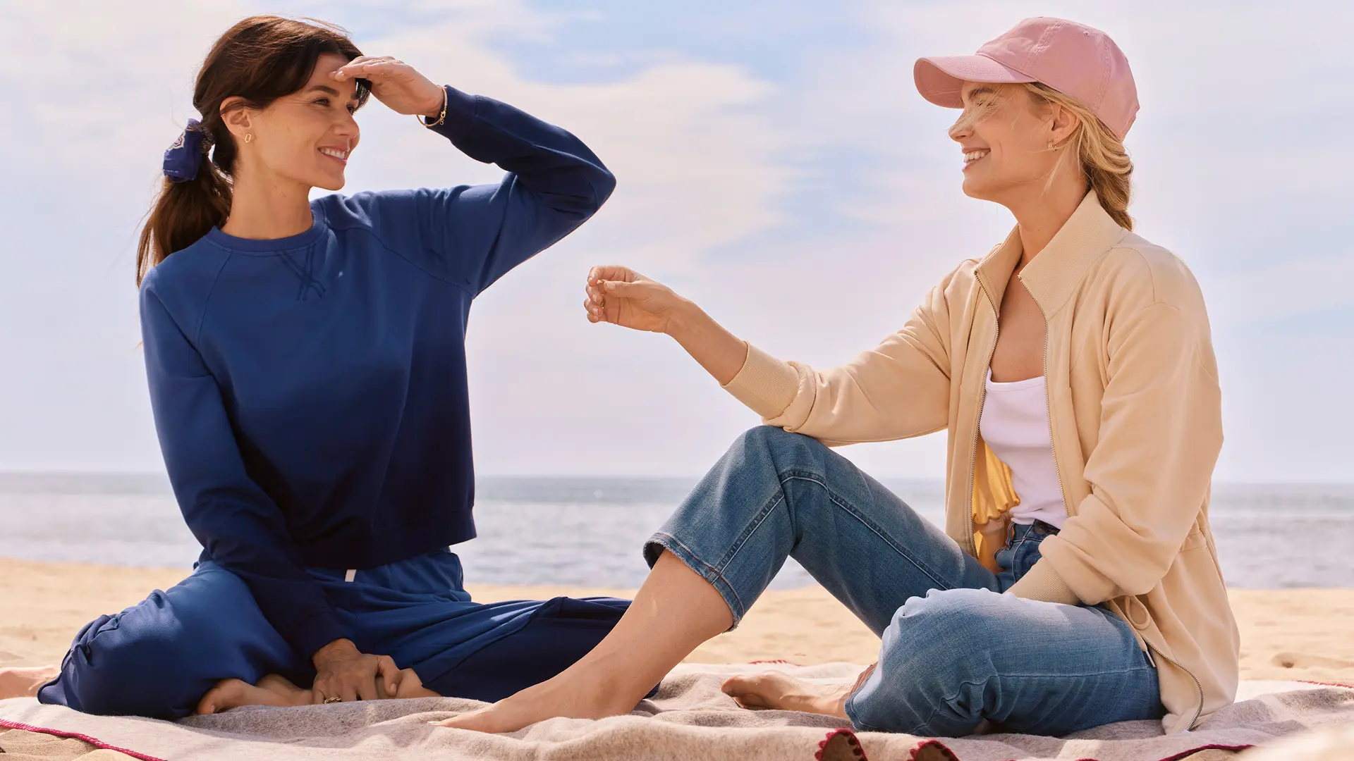 Two women on the beach, one wearing a blue ombre matching sweat set and the other wearing jeans, a creme jacket, and a pink hat